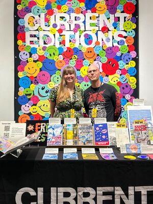 Two people are standing behind a table covered with publications at a vibrant event. The woman on the left is wearing a floral dress, and the man on the right is in a black shirt with red text that reads âINSECT WARFAREâ. Behind them is a colorful, eye-catching backdrop filled with smiley faces in various colors, with the words "CURRENT EDITIONS" prominently displayed at the top.